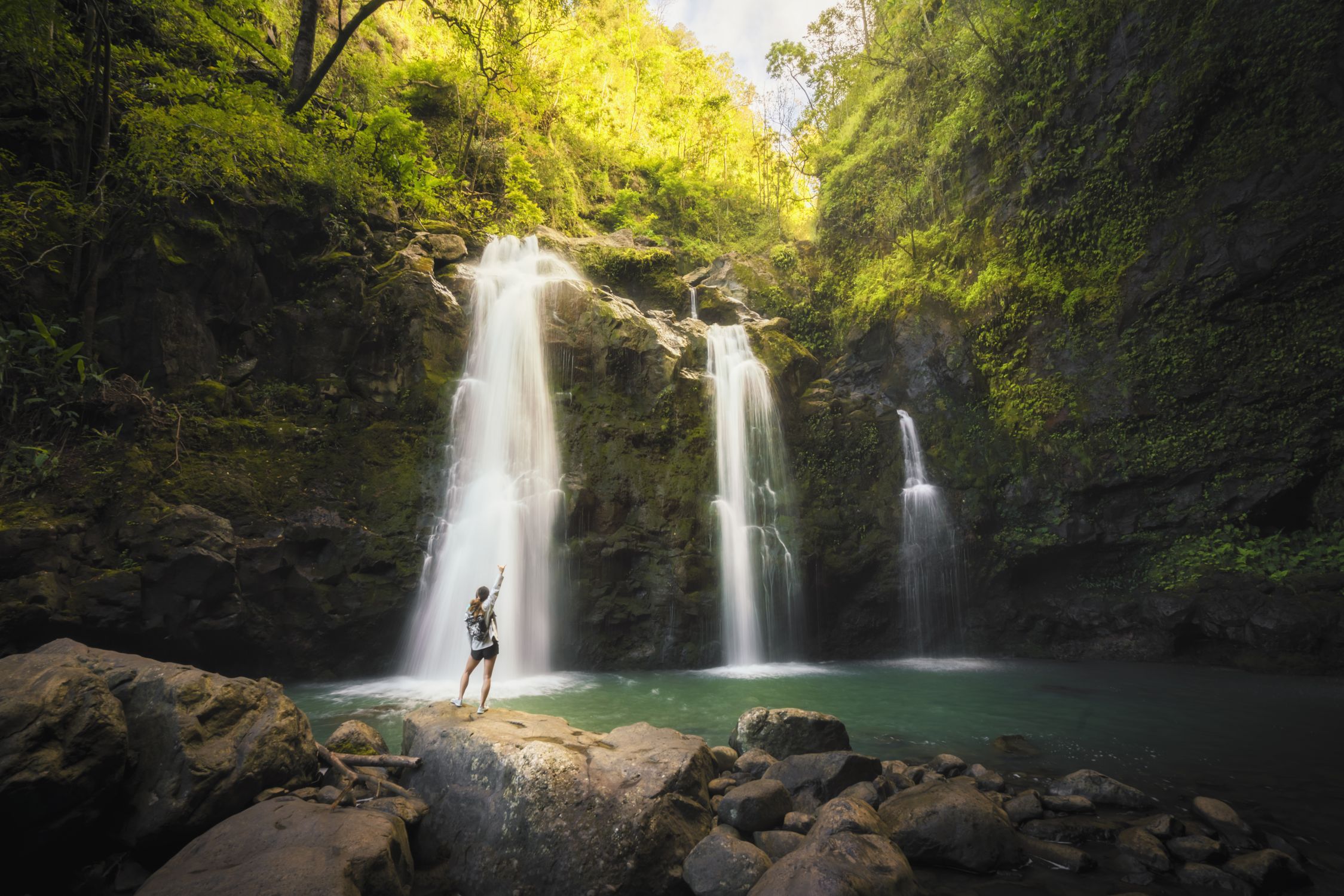 A hiker at the base of a three tier waterfall on Maui